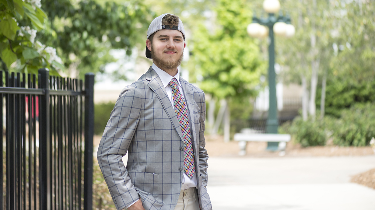 A young man wearing a suit and a baseball cap stands next to an iron fence.