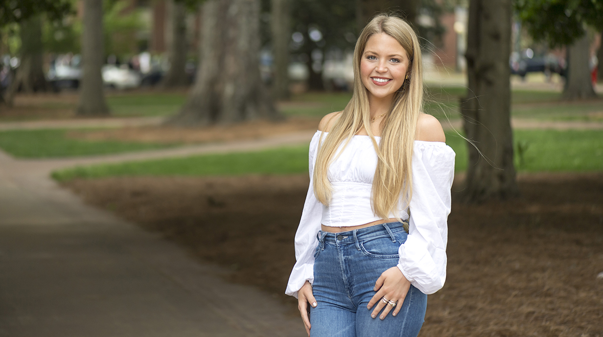 A young lady in jeans stands on a sidewalk in a park.