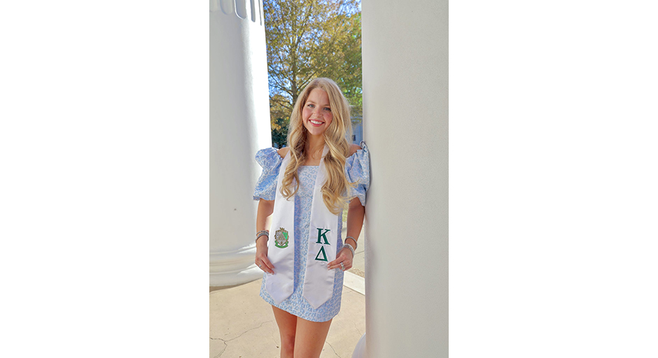 A young woman wearing a stole with Greek letters poses for an outdoor portrait.