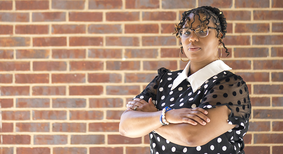 A young woman stands in front of a brick wall with her arms crossed.