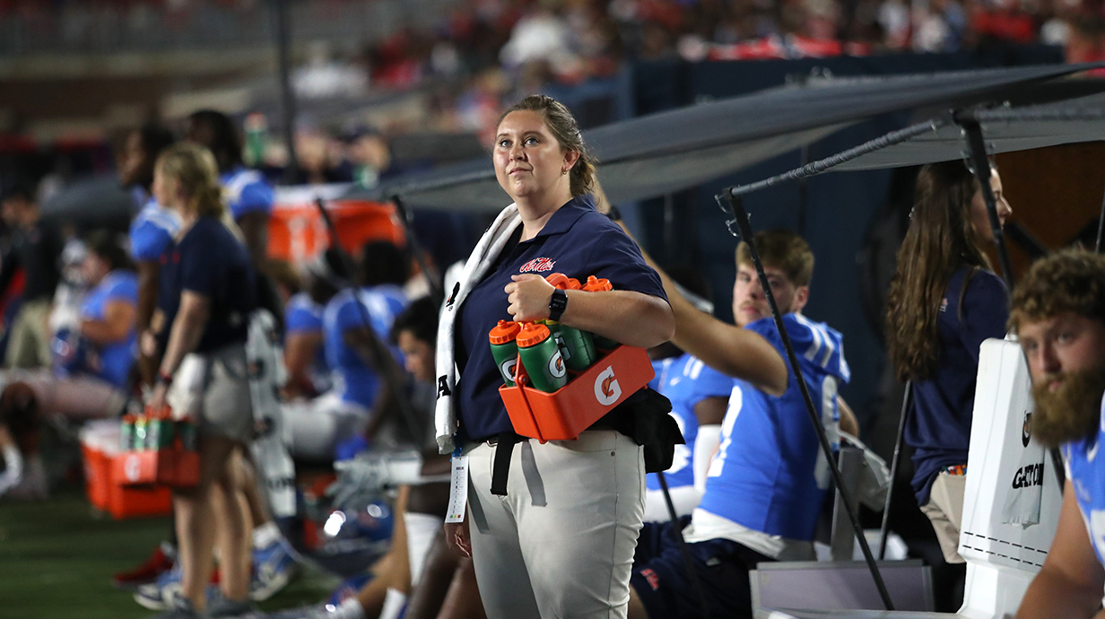 A woman stands on the playing field of a stadium, holding a basket of water bottles.