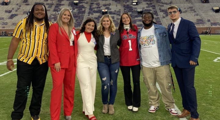 Seven young men and women stand on the playing field of a stadium.