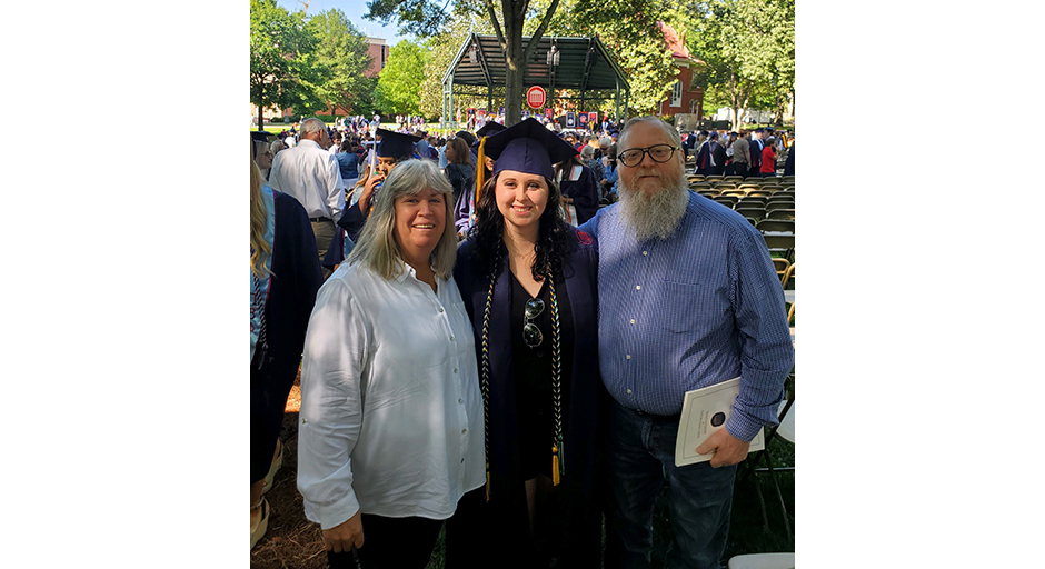 A man and a woman embrace a young woman wearing graduation robes in a park.