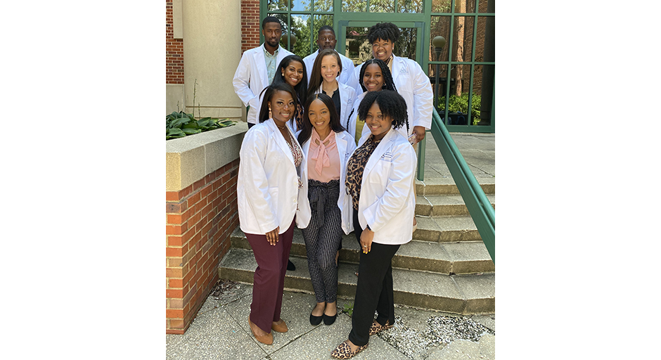 A group of young people wearing white lab coats stand outside on concrete steps.