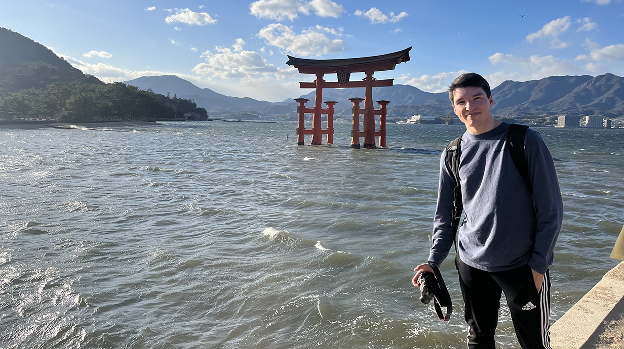 Man holding a camera standing in front of a body of water with a Japanese monument in the background.