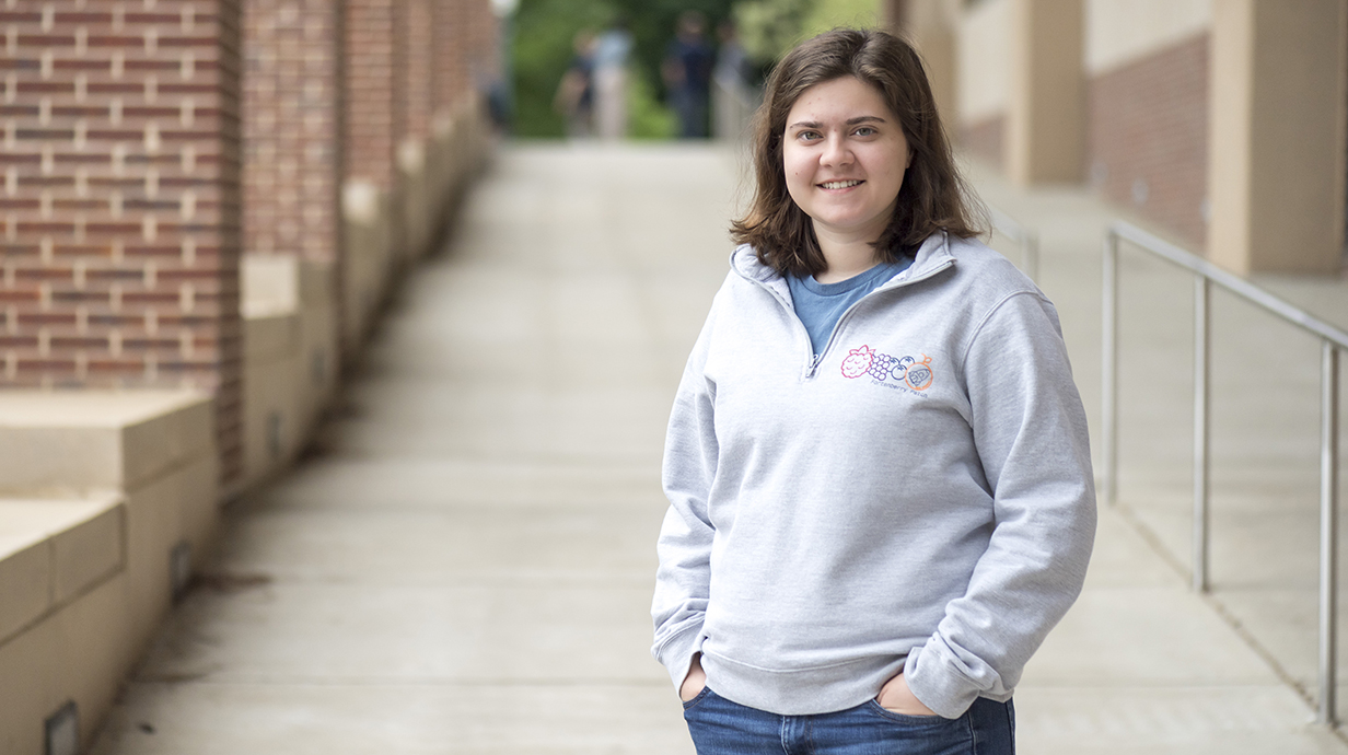 A young woman wearing a gray sweatshirt stands on a covered porch.