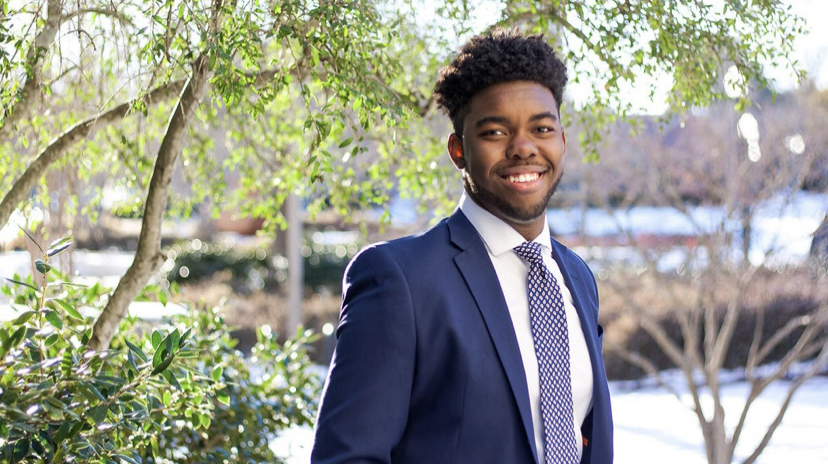A smiling young man in a suit stands in front of some trees.