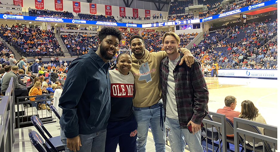 Three young men and a woman pose for a photo in a basketball arena.