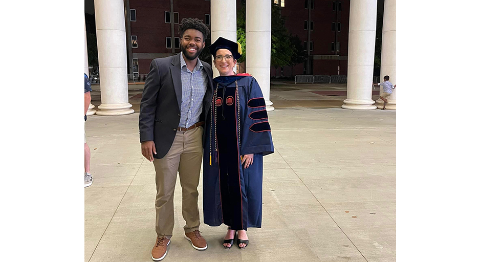 A young man stands next to a woman wearing graduation robes.