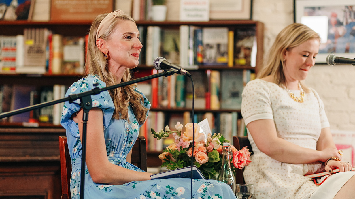 Two women sit on a stage surrounded by bookcases full of books.