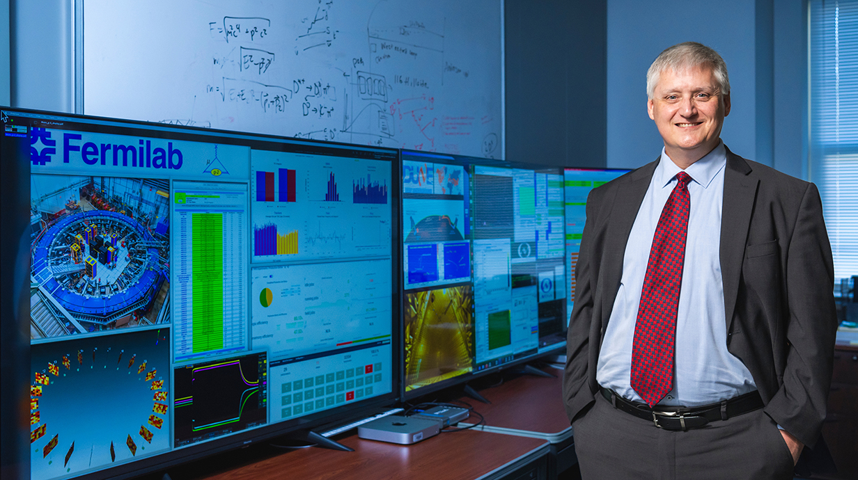 A man wearing a suit stands beside research posters from Fermilab.