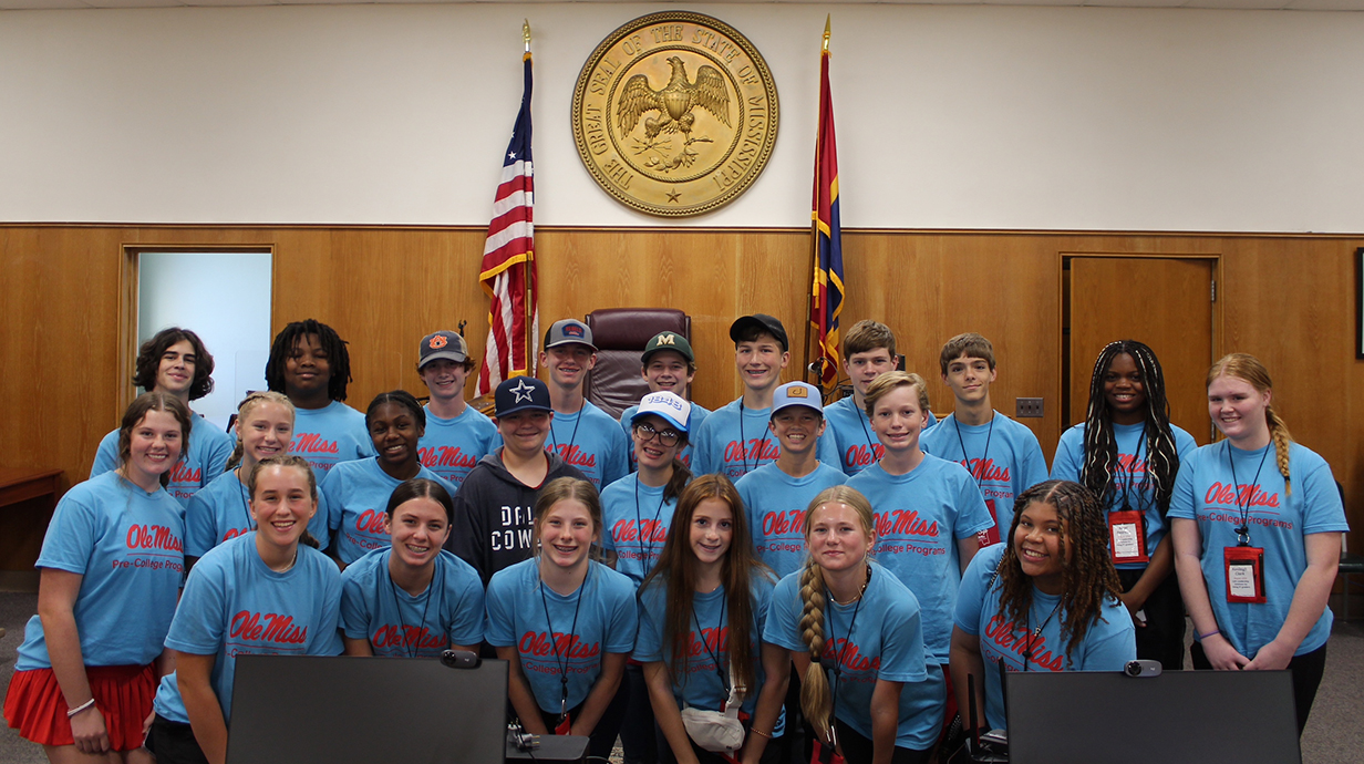 A group of high school students wearing matching blue Ole Miss T-shirts stand in a courtroom in front of the great seal of Mississippi.