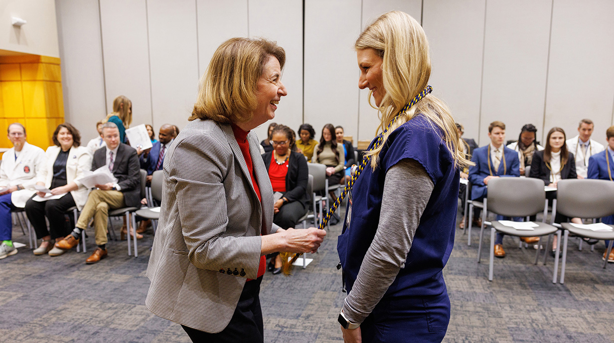 A woman smiles as she presents an honor cord to a young women in front of a crowded conference room.