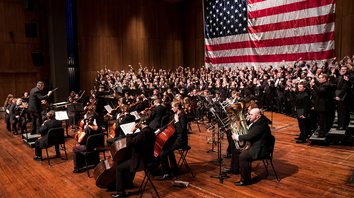 An orchestra performs on a stage in front of a giant American flag.