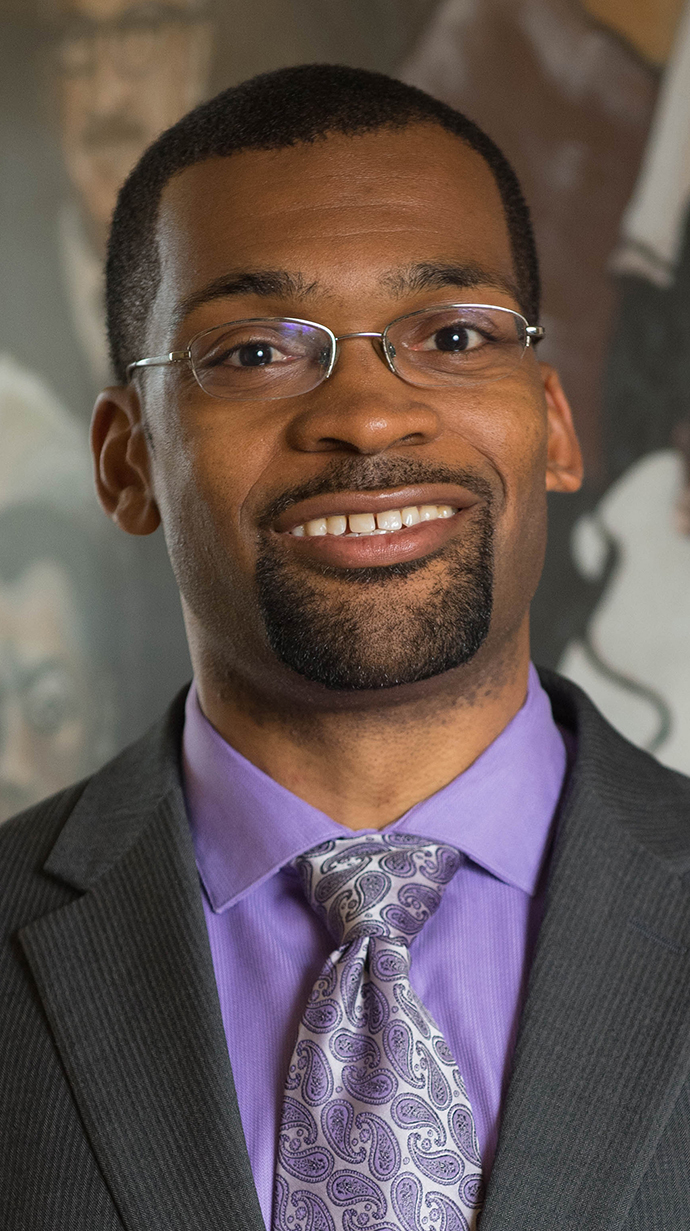 Headshot of a man wearing glasses and a gray suit with a lilac shirt.