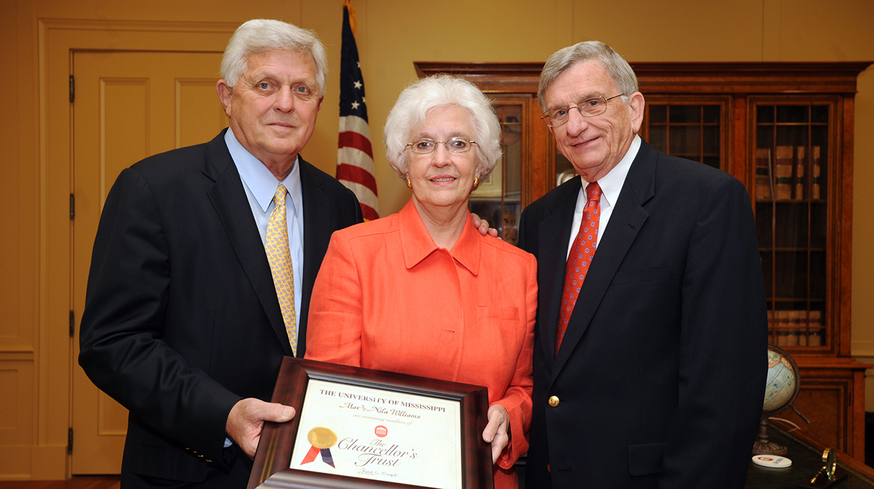 A man wearing a dark suit presents a framed certificate to a woman and a man.