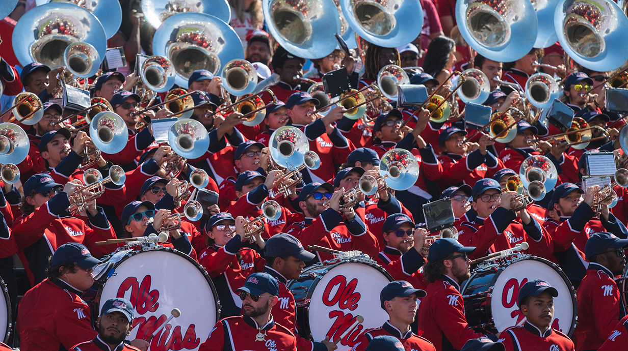 A marching band dressed in bright red and blue uniforms plays in the seating area of a stadium.