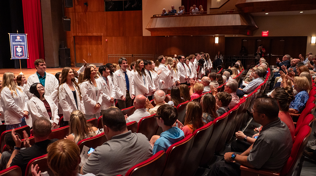 A large group of young people, all wearing white lab coats, stand and face the audience in a crowded auditorium.