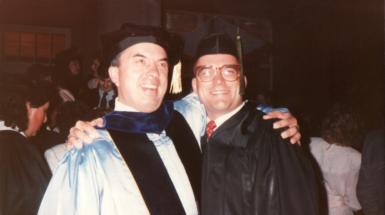 Two men wearing graduation regalia stand with their arms around each other.