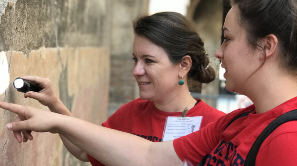 Two women wearing red T-shirts use a flashlight to examine markings on a stone wall.