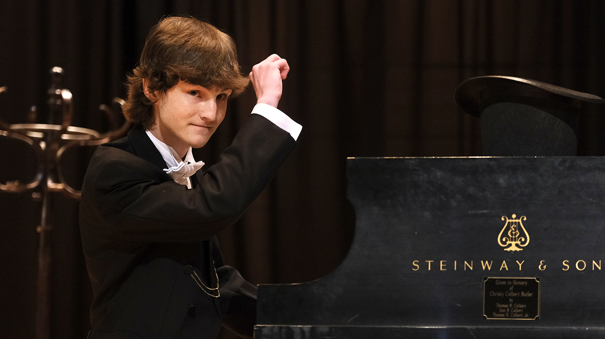 A young man wearing a tuxedo plays a piano on a stage.