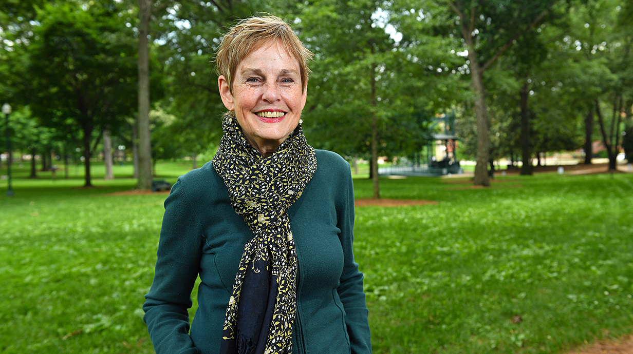 A woman wearing a large patterned scarf stands in a tree-lined park.