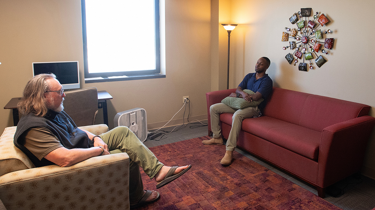 A man sitting in an upholstered chair talks with a young man seated across from him on a sofa.