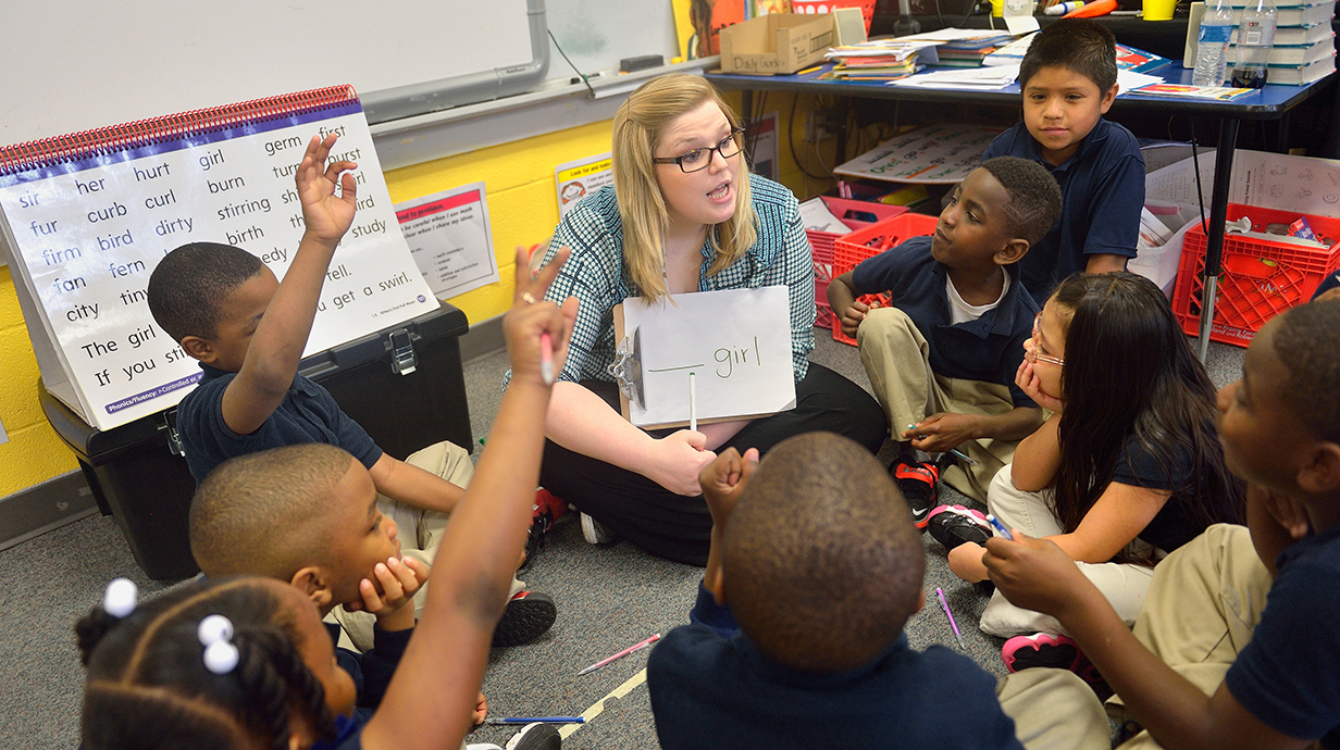 A teacher sits in the floor of a classroom with a group of young children to teach them reading.