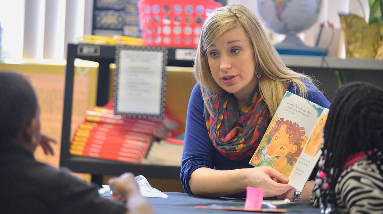 A woman holds a picture book open in front of two young children in a classroom.