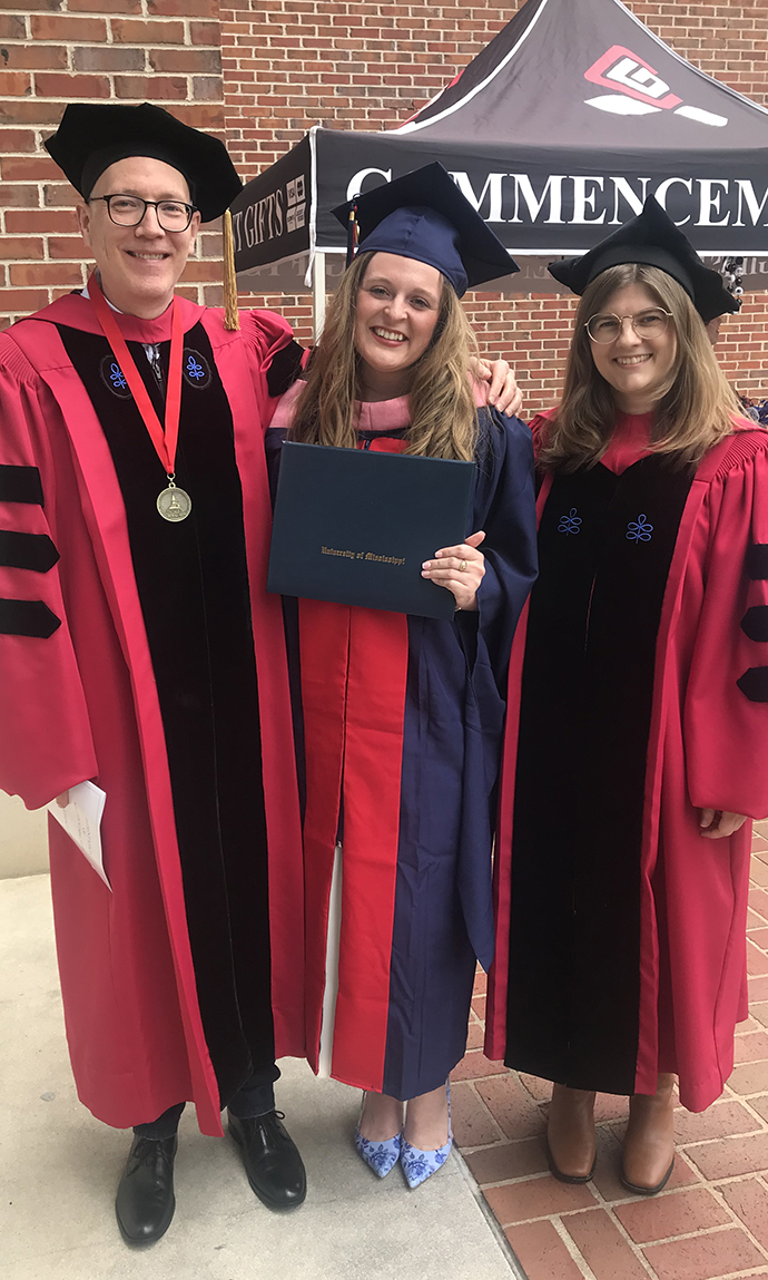 A man and two women, all wearing red graduation robes, stand in front of a pop-up tent reading 'Commencement.'