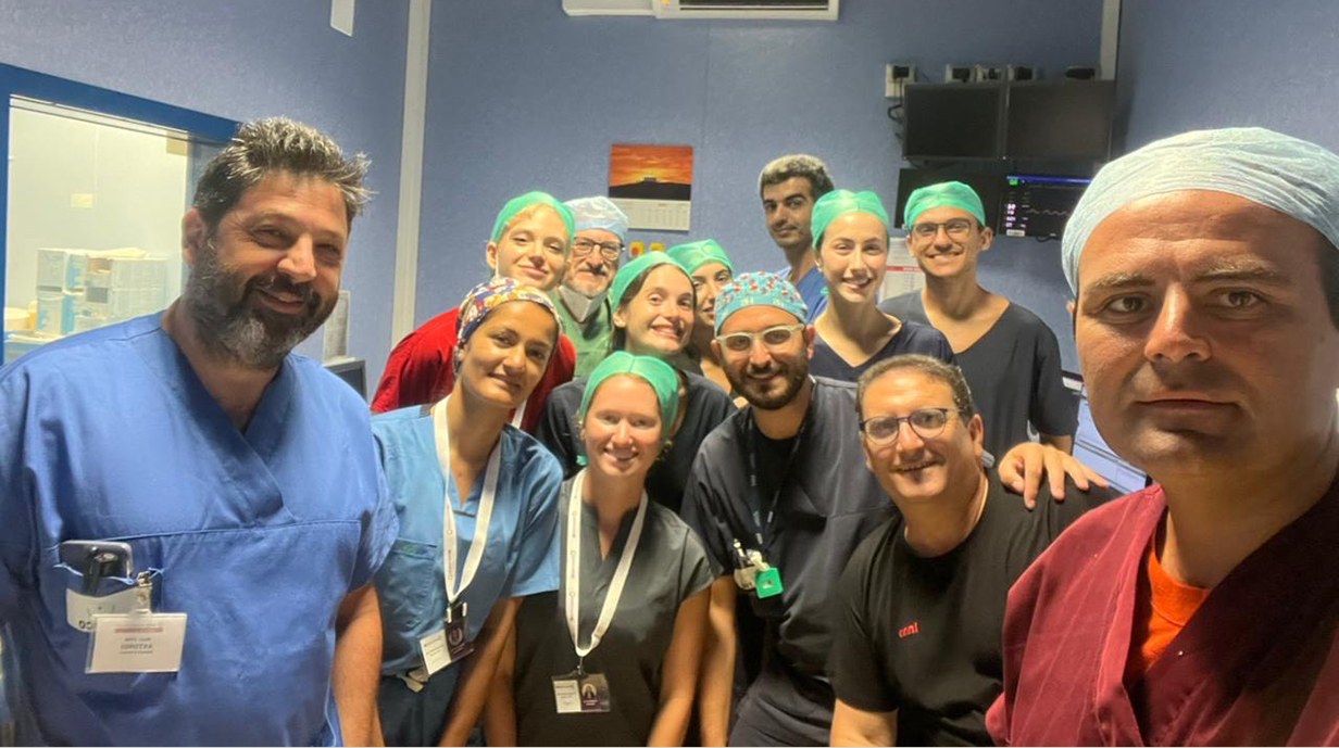 A group of people, all wearing surgical scrubs, gather for a photo outside an operating room.