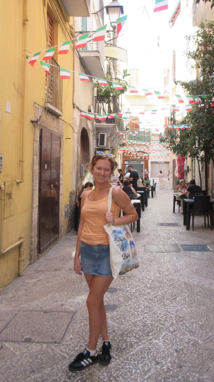 A young woman stands on a cobblestone street lined with tables and colorful flags in Italy.