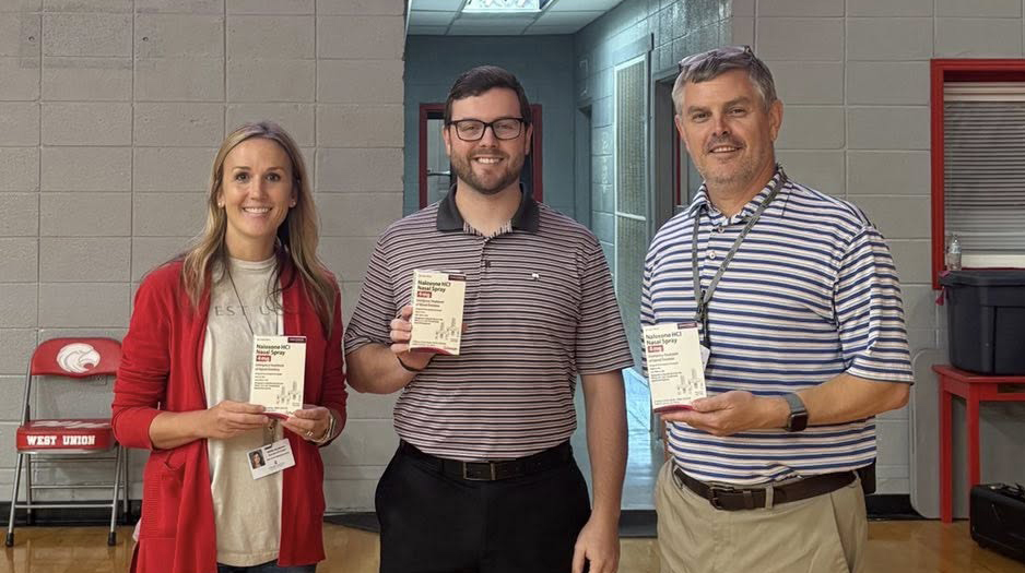 A woman and two men stand in a high school gym holding boxes of product.