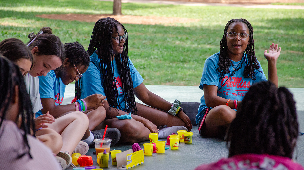 A teenage girl sitting outside at the end of a line of girls raises her hand.