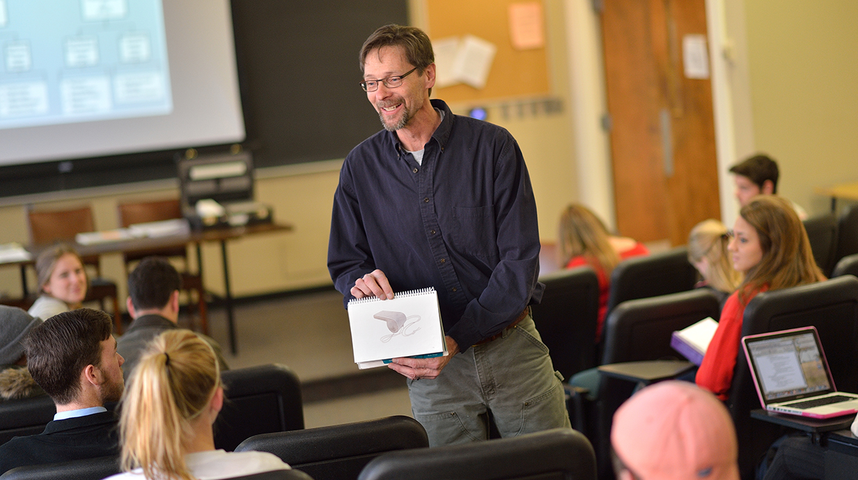 A smiling teacher shows a drawing of a whistle to students in a college classroom.