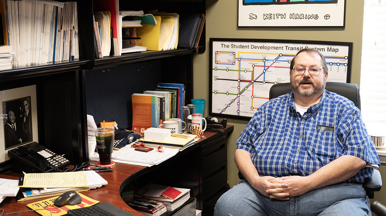 A man sits at an L-shaped desk lined with books, folders and memorabilia.