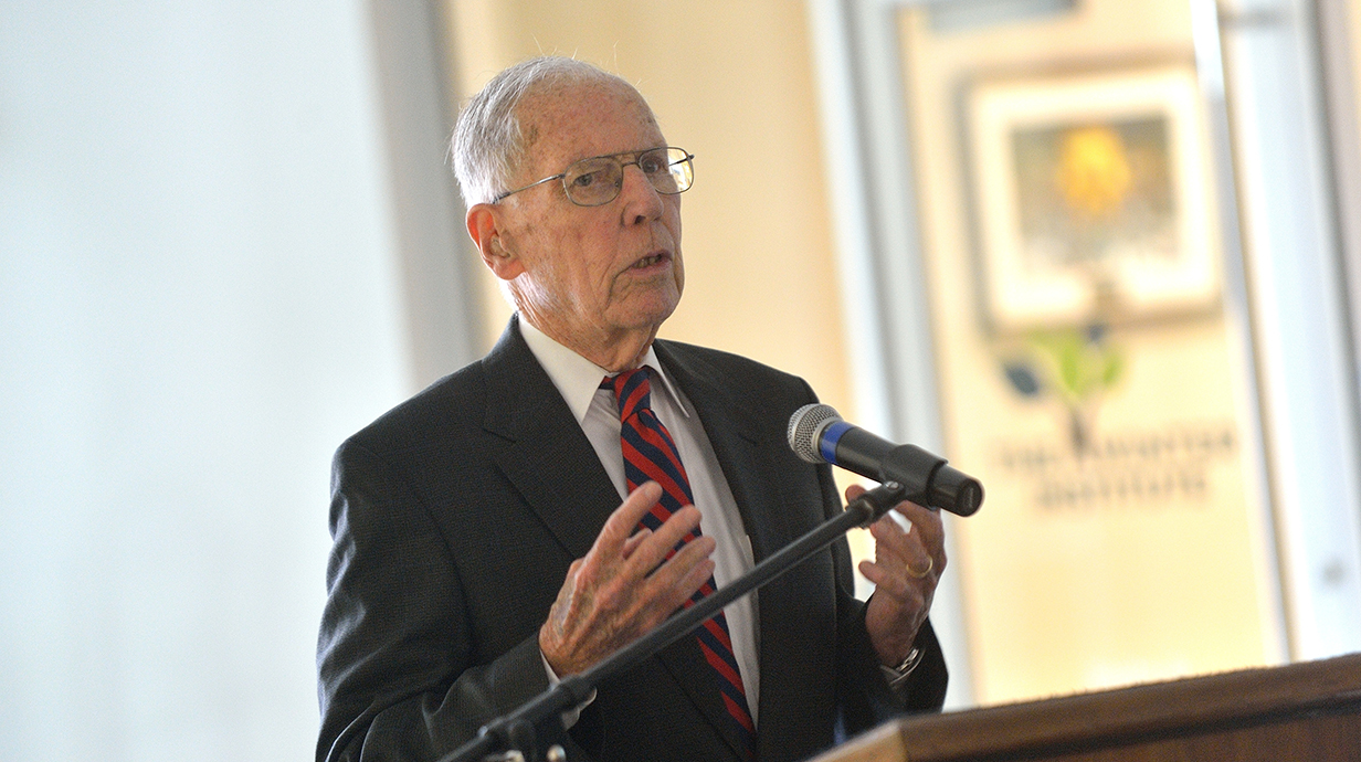 An older man wearing a suit speaks at a podium.