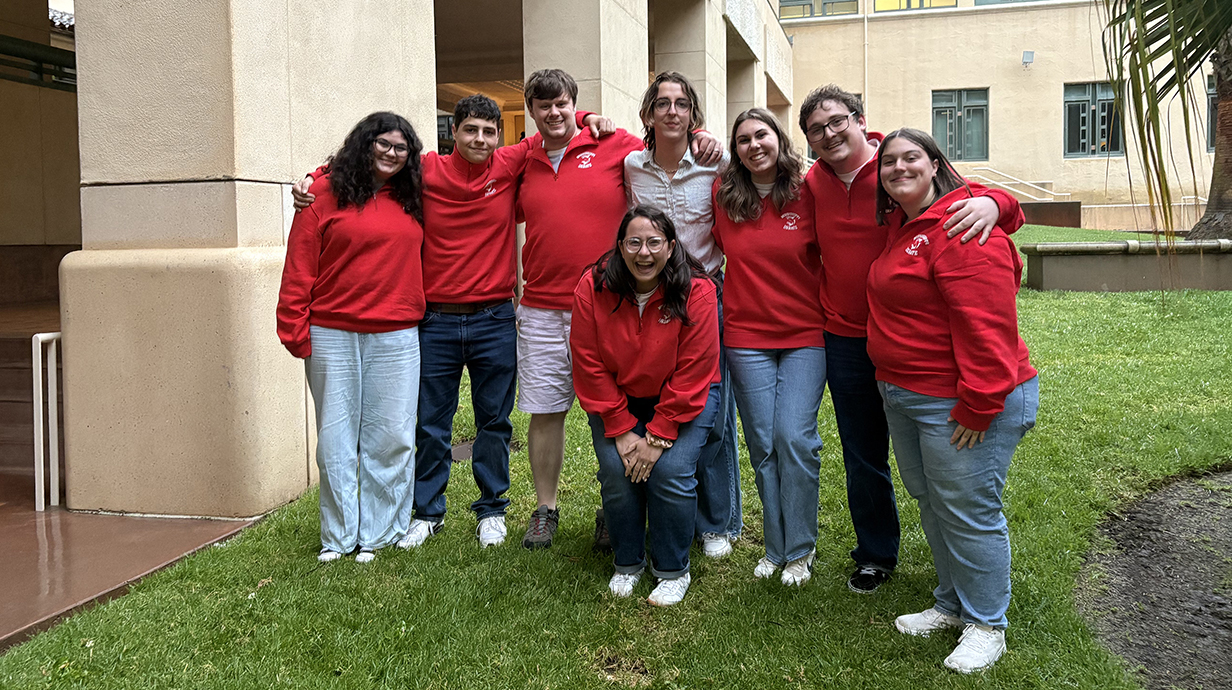 A group of young people, most wearing matching red sweatshirts, stand in a grassy area outside a large stucco building.