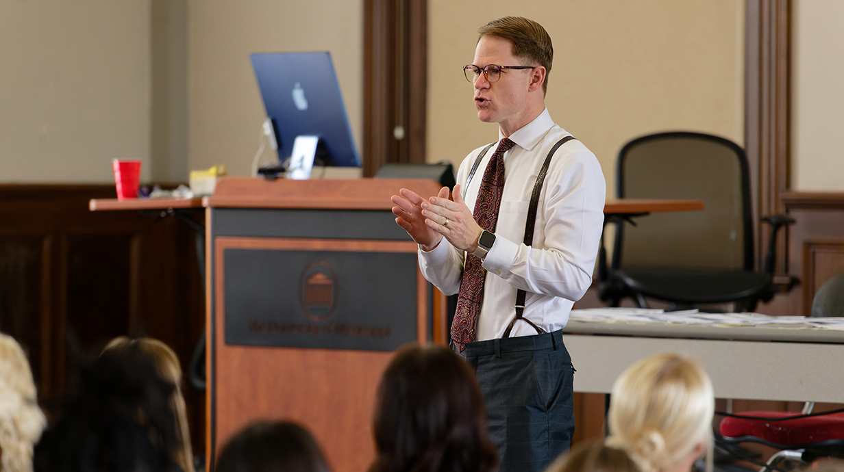 A man wearing a necktie and suspenders speaks in a classroom to a group of students.