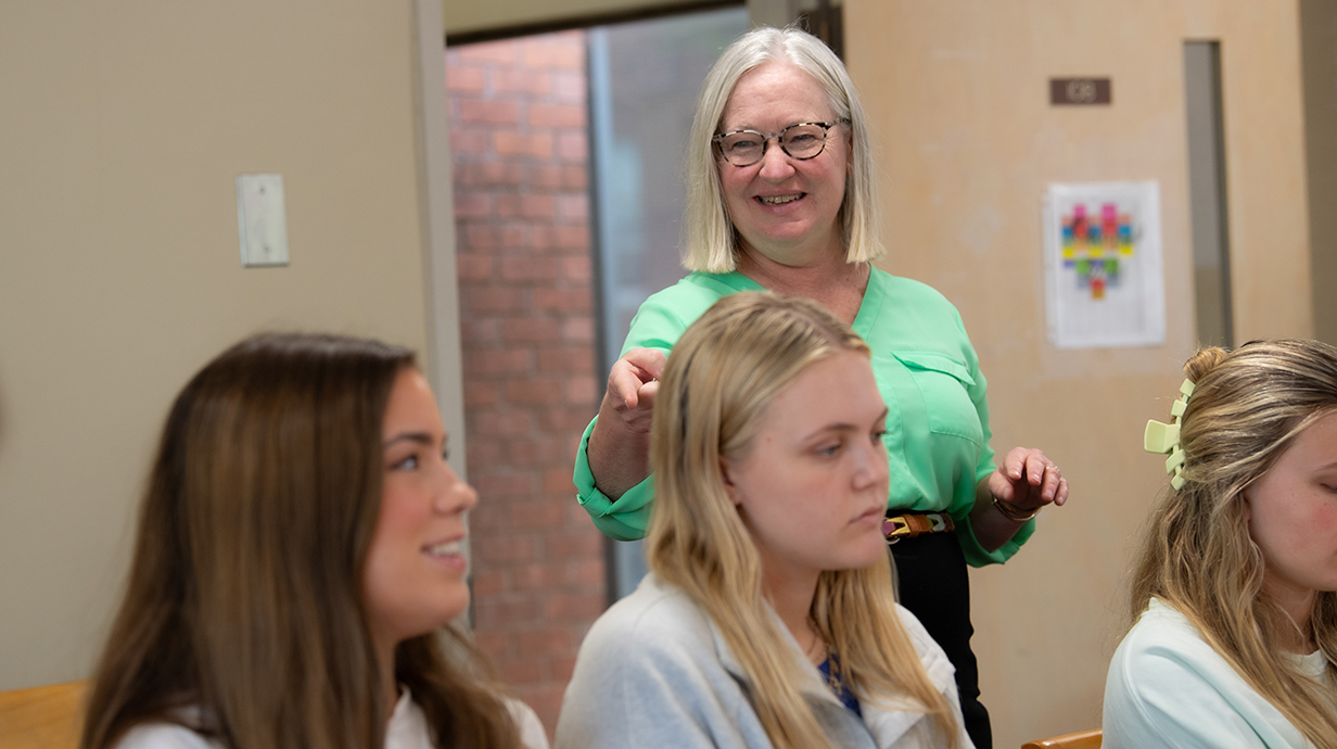 A woman wearing a green blouse points at a smiling young women while lecturing in a crowded classroom.