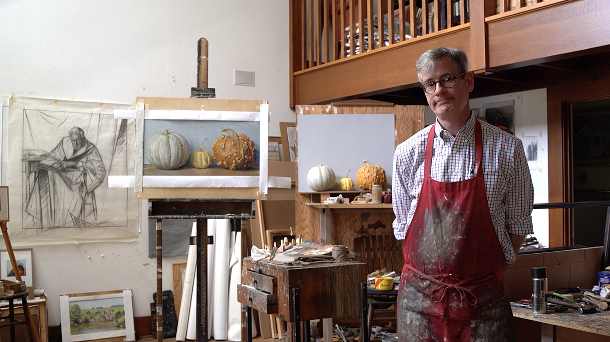 A man wearing a red apron stands in a painter's studio with artwork lining the walls.