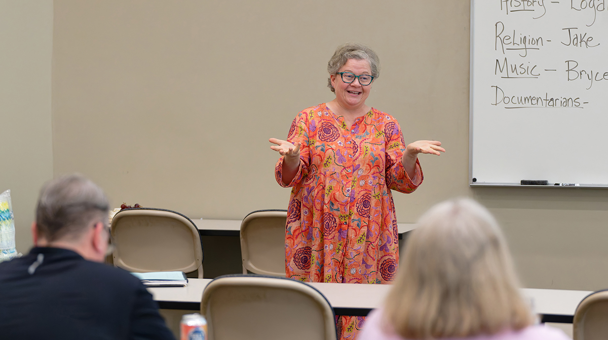 A woman wearing an orange dress speaks to a group from the front of a classroom.