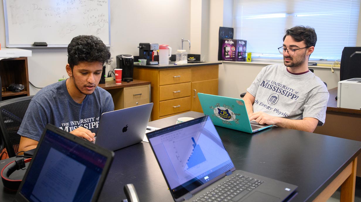 Two young men at a table with their laptops open. 