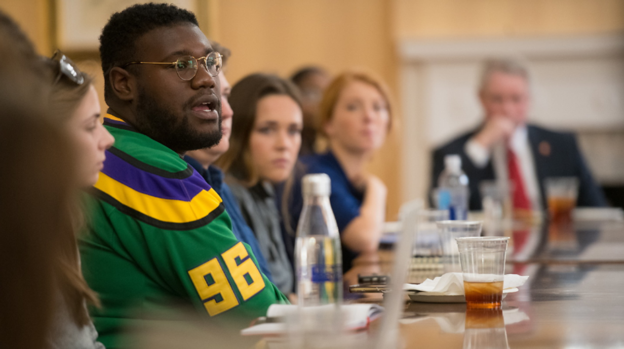 A diverse group of students participating in student affairs sits with the university chancellor at a long conference table. 