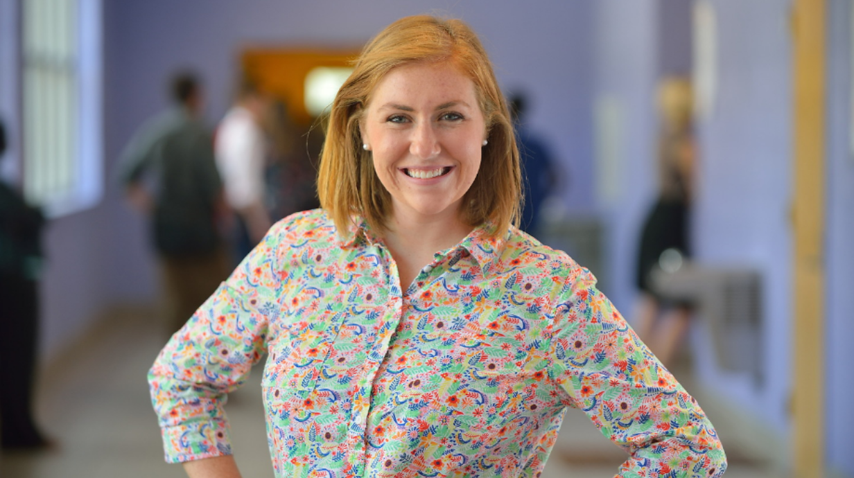 Teacher stands in school hallway smiling. Students appear in the background.