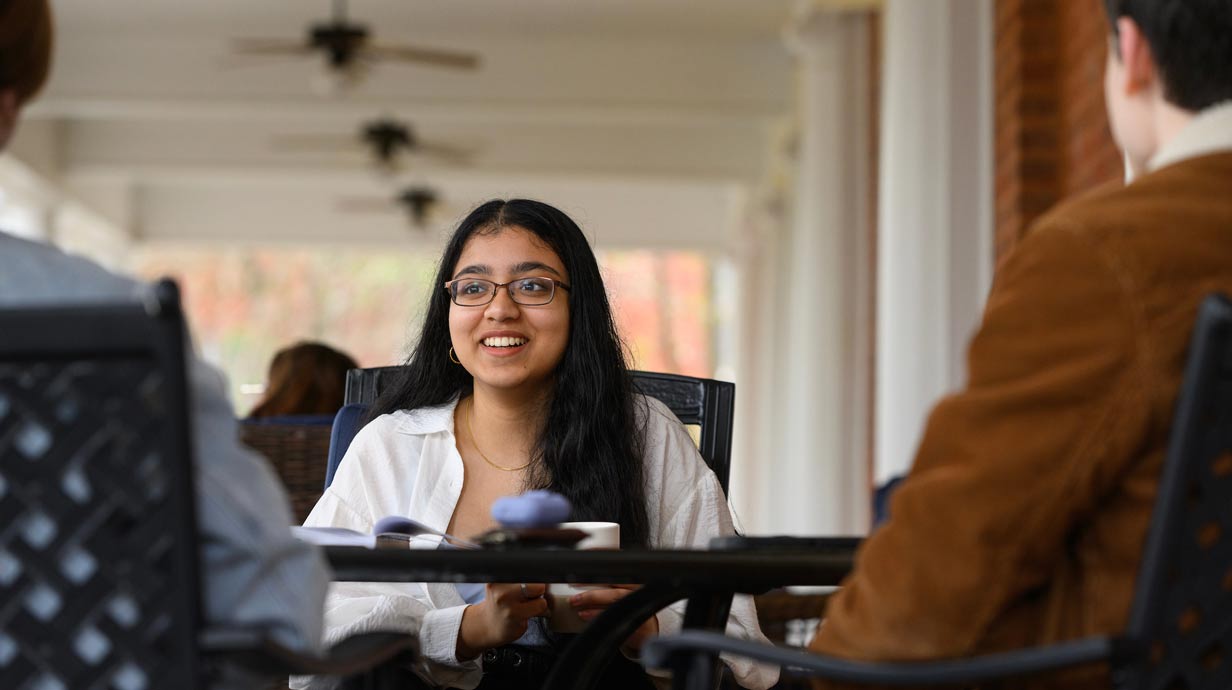 Croft student, Aahba Mantri, drinking coffee with two other students on the porch of the Croft building