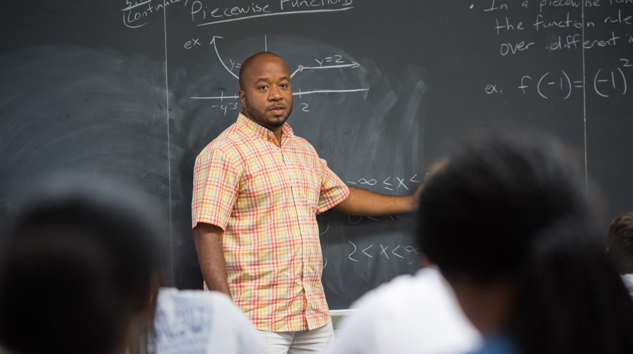 Professor at chalkboard gesticulating with hands and turning to class.