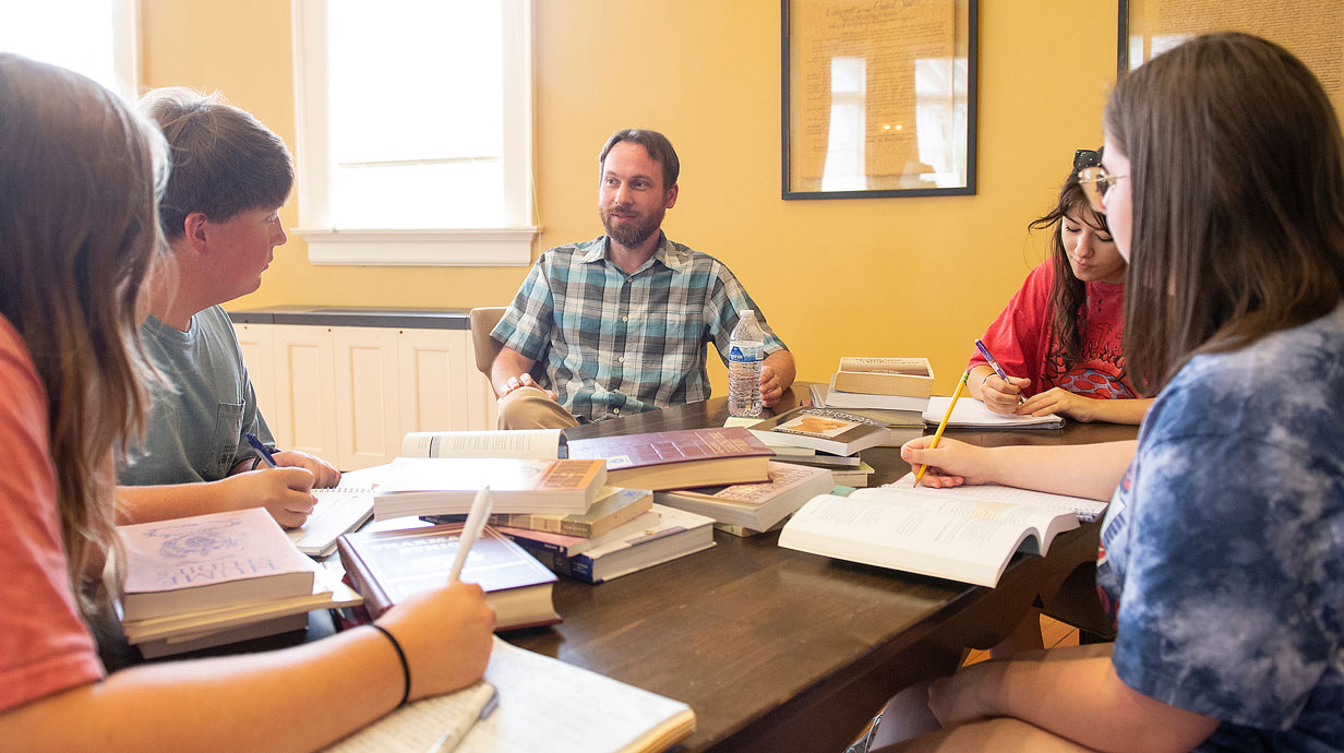 Students sit around a table listening to a professor. 