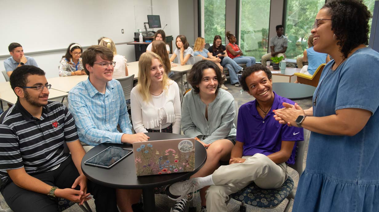 Group of students seated talking to a teacher that is standing