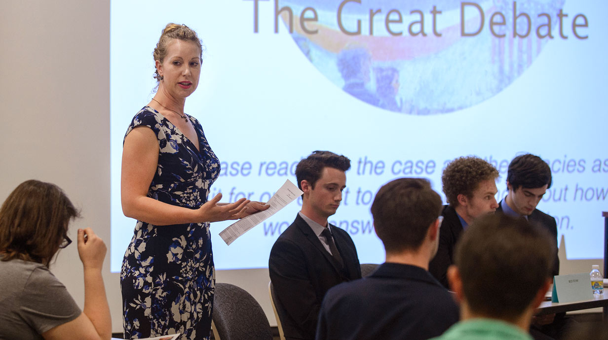 Professor holding a sheet of paper in front of a screen that says "The Great Debate" surrounded by professionally dressed students sitting at tables.